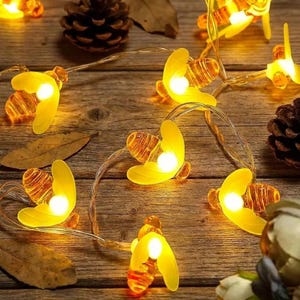 May include: A string of yellow bee-shaped fairy lights with a warm white glow. The lights are strung on a thin, silver wire and are scattered on a wooden surface with pine cones and leaves.