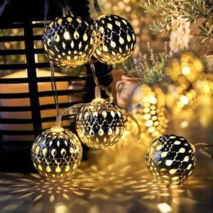 May include: Decorative string lights with spherical, patterned metal shades. The lights are warm yellow and cast intricate shadows. The background includes a black lantern, plants, and a blurred, soft focus effect, creating a cozy ambiance.