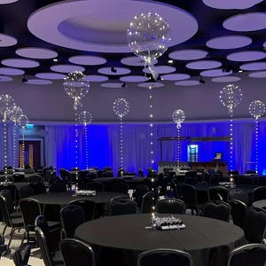 May include: A large event space decorated for a celebration. Clear balloons filled with small white lights float above round black tables. The ceiling has circular white accents and the walls are draped in blue fabric.