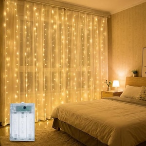 Op de afbeelding: Warme witte lichtslingers verlichten transparante gordijnen in een slaapkamer. Een batterijpack is zichtbaar op de voorgrond. De kamer heeft een bed met een beige dekbed en een nachtkastje met een lamp.