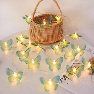 Könnte beinhalten: Eine Lichterkette mit schmetterlingsförmigen Lichtern in Blau und Grün, die warm leuchten. Im Hintergrund ist ein geflochtener Korb zu sehen.