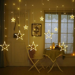 May include: A decorative display of star-shaped fairy lights hanging from the ceiling. The warm white lights illuminate the star outlines, creating a cosy atmosphere. A small table and chair are also visible in the scene.