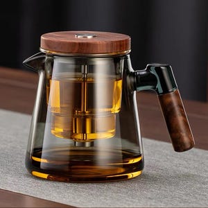 May include: A glass teapot with a wooden lid and handle. The teapot has a built-in infuser and is filled with amber-colored liquid. The handle is dark brown, and the lid is a lighter wood tone. The teapot sits on a gray surface.