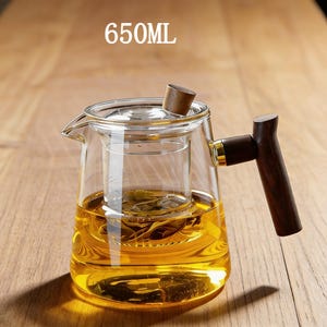 May include: A clear glass teapot with a 650ml capacity, featuring a removable glass infuser containing tea leaves, a wooden lid knob, and a dark brown wooden side handle with a gold-colored metal accent connecting it to the glass body.