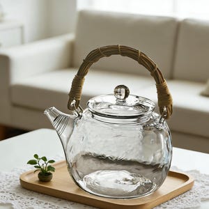 Könnte beinhalten: Eine klare Glas-Teekanne mit strukturierter Oberfläche und einem geflochtenen Griff. Die Teekanne hat einen Glasdeckel mit einem kleinen Knopf und einen Ausguss mit vertikalen Linien. Sie steht auf einem Holztablett.