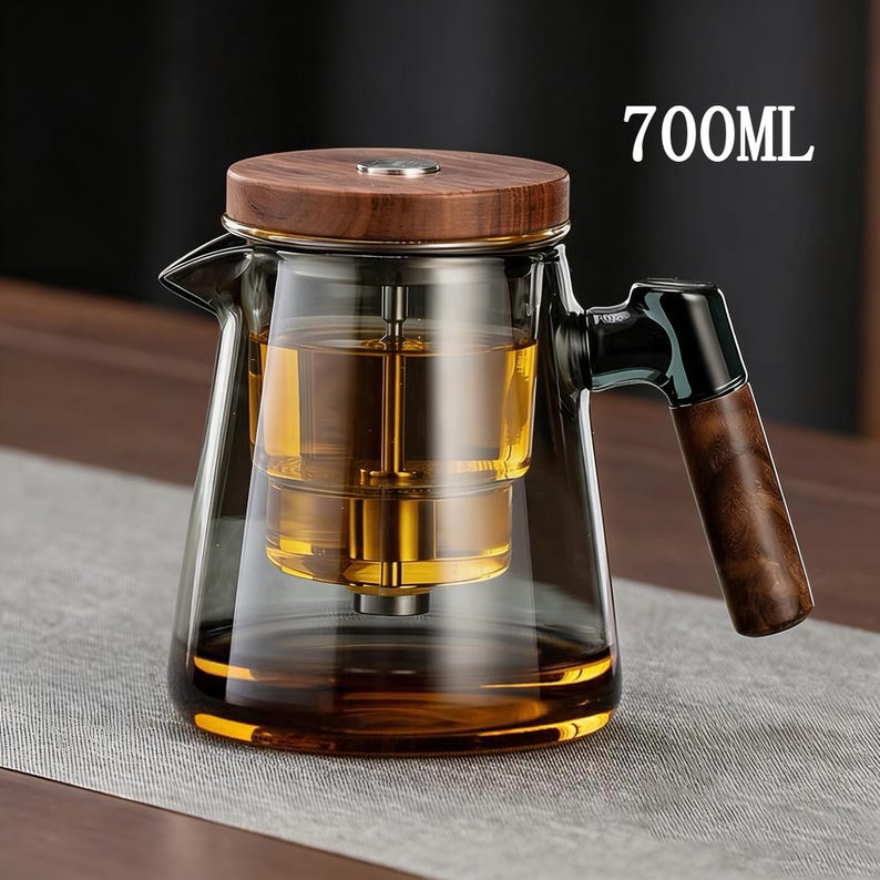 Pode incluir: Uma chaleira de vidro transparente com tampa e al&ccedil;a de madeira. A chaleira tem um infusor embutido e &eacute; preenchida com l&iacute;quido &acirc;mbar. O texto "700ML" &eacute; vis&iacute;vel no lado direito da chaleira.