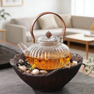 May include: A clear glass teapot with a wooden handle and lid, filled with tea. The teapot sits in a dark brown, textured bowl filled with stones. The teapot has a ribbed design and is on a table.