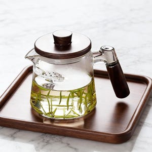 May include: A glass teapot with a wooden lid and handle, containing tea and herbs. The teapot is on a wooden tray. The handle is dark wood and stainless steel. The tea is a pale yellow colour. The tray is approximately 25 cm long.