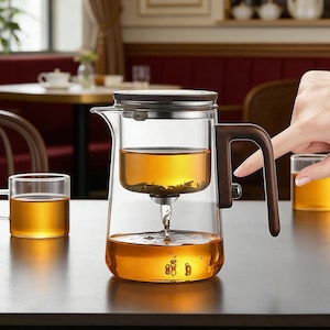 May include: A clear glass tea infuser with a dark wooden handle and lid. The infuser is filled with tea and is pouring tea into the main chamber. A clear glass mug of tea sits to the left. The background includes a table and chairs.