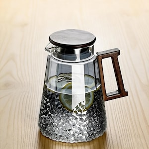 May include: A clear glass pitcher with a wooden handle and lid. The pitcher is filled with water and lemon slices. The bottom of the pitcher has a textured diamond pattern. The handle and lid are made of dark wood.