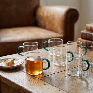 May include: A set of clear glass mugs with emerald green handles arranged on a wooden surface. One mug is filled with amber-colored tea, and a small white plate with a piece of biscotti sits nearby. The mugs feature a simple, cylindrical design.
