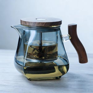 Peut inclure: Une théière en verre avec un couvercle et une poignée en bois. La théière a un corps en verre transparent avec une teinte bleue et un infuseur en acier inoxydable. La poignée est en bois foncé. La théière est remplie de thé.
