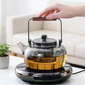 May include: A clear glass teapot with a dark wooden handle and a black metal frame, filled with amber liquid. The teapot sits on a black electric warmer. A small potted plant is to the left.