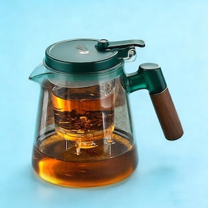 Op de afbeelding: Een doorzichtige glazen theepot met een donkergroen deksel en handvat, en een houten handgreep. De theepot bevat gezette thee en een thee-infuser. De achtergrond is lichtblauw.