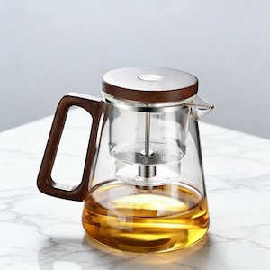 Pode incluir: Uma chaleira de vidro transparente com uma alça e tampa de madeira. A chaleira tem um infusor de aço inoxidável e é preenchida com líquido de cor âmbar. A alça e a tampa são feitas de madeira escura.