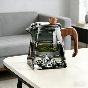 May include: A clear glass teapot with a wooden handle and lid. The teapot has a built-in infuser and a mountain-shaped design at the base. The teapot is filled with green tea and sits on a white table.