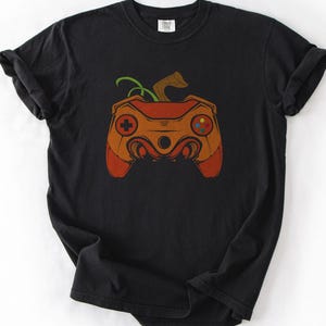 May include: A black short-sleeve t-shirt featuring a graphic print of an orange pumpkin shaped like a video game controller. The controller design includes a green stem, a directional pad, and colorful buttons on a dark background.