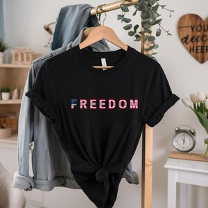 May include: Black t-shirt with the word "FREEDOM" in a red, white, and blue design. The shirt is knotted at the waist and hangs on a wooden hanger. A denim jacket hangs behind it.