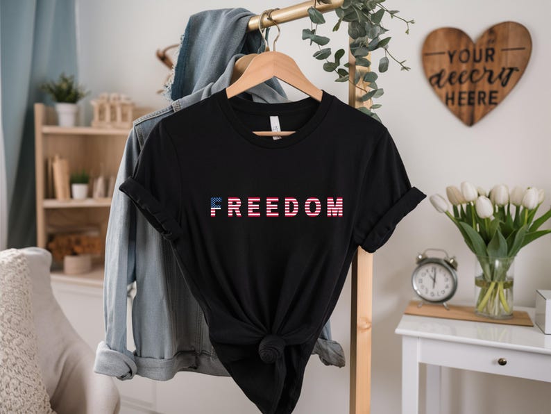 May include: Black t-shirt with the word "FREEDOM" in a red, white, and blue design. The shirt is knotted at the waist and hangs on a wooden hanger. A denim jacket hangs behind it.