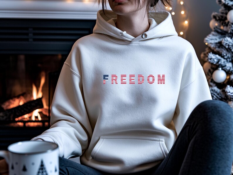 May include: Cream-colored hoodie with the word "FREEDOM" across the chest in a red, white, and blue American flag design. The hoodie has a front pocket and a drawstring hood. The image is set in front of a fireplace.