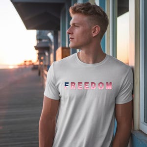 May include: White t-shirt with the word "FREEDOM" printed across the chest in red, white, and blue. The design incorporates the colors of the American flag. The shirt is short-sleeved and made of a soft material.