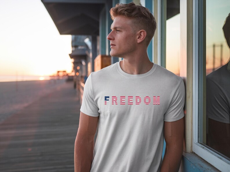May include: White t-shirt with the word "FREEDOM" printed across the chest in red, white, and blue. The design incorporates the colors of the American flag. The shirt is short-sleeved and made of a soft material.