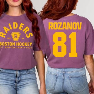 May include: A purple t-shirt with the text "RAIDERS BOSTON HOCKEY" in yellow. The back of the shirt displays "ROZANOV 81" in yellow. The shirt is a casual style, suitable for sports fans.