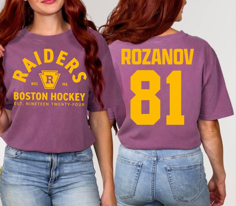 May include: A purple t-shirt with the text "RAIDERS BOSTON HOCKEY" in yellow. The back of the shirt displays "ROZANOV 81" in yellow. The shirt is a casual style, suitable for sports fans.