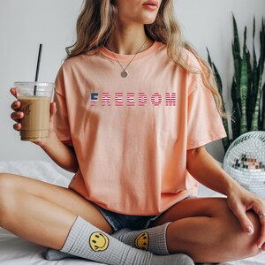 May include: A peach-colored t-shirt with the word "FREEDOM" printed across the chest in red, white, and blue. The shirt is worn with denim shorts and gray socks with yellow smiley faces. A clear cup of coffee is held in the hand.
