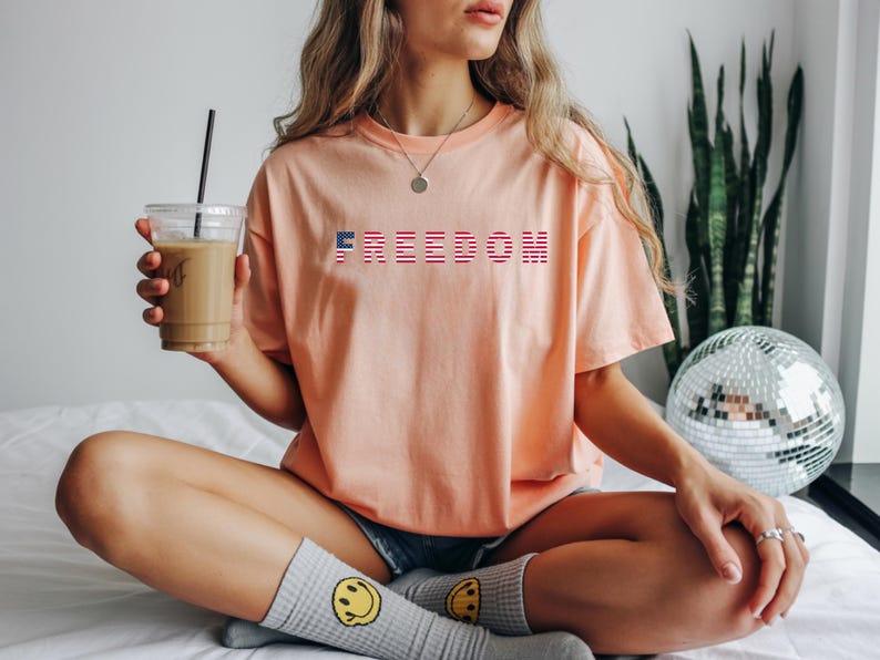 May include: A peach-colored t-shirt with the word "FREEDOM" printed across the chest in red, white, and blue. The shirt is worn with denim shorts and gray socks with yellow smiley faces. A clear cup of coffee is held in the hand.