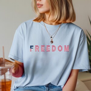 May include: Light blue t-shirt with the word "FREEDOM" printed across the chest. The letters are designed with a red and white striped pattern, and the letter "F" is a blue field of stars. The shirt is a casual style.