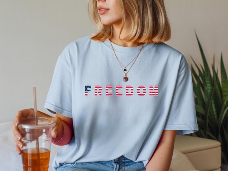 May include: Light blue t-shirt with the word "FREEDOM" printed across the chest. The letters are designed with a red and white striped pattern, and the letter "F" is a blue field of stars. The shirt is a casual style.