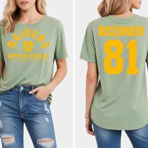 May include: Sage green t-shirt with the word "RAIDERS" arched across the chest in yellow, along with a logo and "BOSTON HOCKEY" text. The back of the shirt displays "ROZANOV" and the number "81" in yellow.