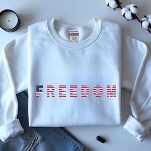 May include: A white sweatshirt with the word "FREEDOM" printed across the front in red and blue, with the letter "F" in a flag design. The sweatshirt is folded on top of a pair of blue jeans.