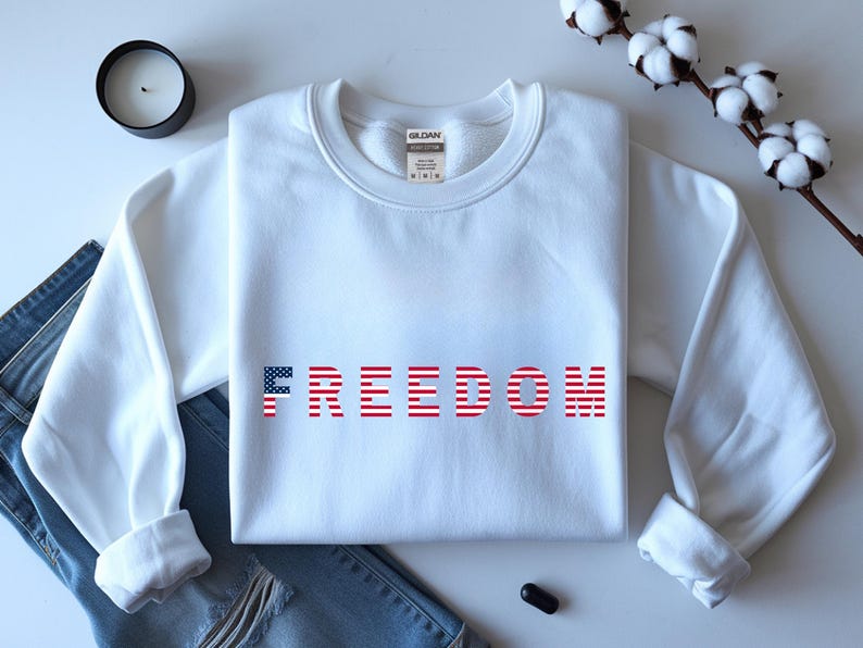May include: A white sweatshirt with the word "FREEDOM" printed across the front in red and blue, with the letter "F" in a flag design. The sweatshirt is folded on top of a pair of blue jeans.
