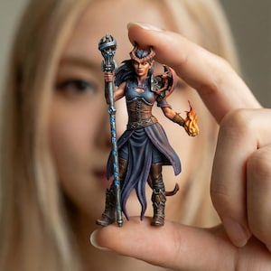 Peut inclure: Figurine fantastique miniature d'un personnage féminin avec des cornes, tenant un bâton et une main enflammée. La figurine est vêtue d'une armure bleue et marron avec un dragon sur l'épaule. La figurine est tenue entre deux doigts.