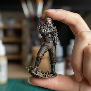 Puede incluir: Figura en miniatura de un guerrero con cabello plateado, vestido con armadura de cuero marrón y cota de malla, sosteniendo dos espadas. La figura se encuentra sobre una pequeña base con hierba y setas. La figura se sostiene entre dos dedos.