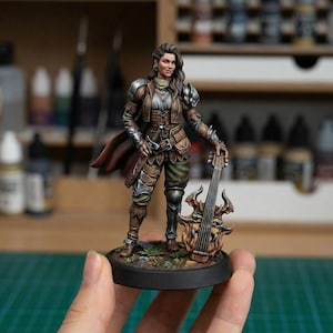 Pode incluir: Figura em miniatura de uma personagem feminina em traje de fantasia, segurando um instrumento de cordas. A figura é pintada com armadura detalhada, uma jaqueta de couro marrom e uma capa. O instrumento tem um design ornamentado.