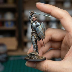 Może przedstawiać: Miniaturowa figurka rycerza w srebrnej zbroi, trzymającego miecz w górze i tarczę z herbem. Rycerz stoi na skalistej podstawie. Figurka jest trzymana w dłoni, ukazując szczegóły malowania.