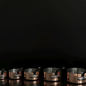 Puede incluir: Un conjunto de cinco cacerolas de color cobre con asas negras, dispuestas en fila sobre un fondo oscuro. Las cacerolas varían en tamaño, con un interior brillante y reflectante.