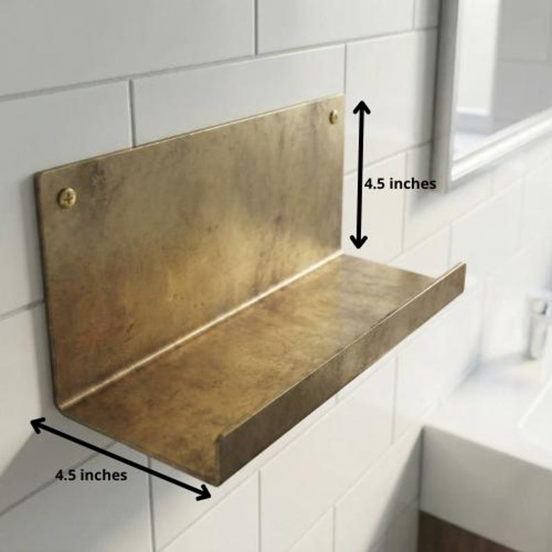 May include: A brass-colored metal shelf, 4.5 inches wide and 4.5 inches high, mounted on a white tiled wall. The shelf has a simple, minimalist design with a small lip to prevent items from sliding off.