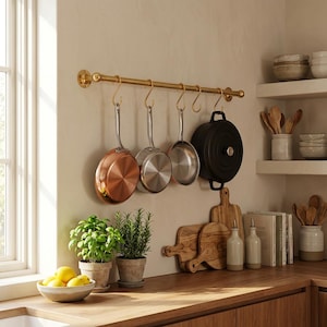 May include: A kitchen scene featuring a brass pot rack with hanging cookware. Copper and stainless steel pans, and a black cast iron pot are suspended. Wooden cutting boards, potted herbs, and bowls of lemons sit on a wooden countertop, creating a warm, inviting atmosphere.