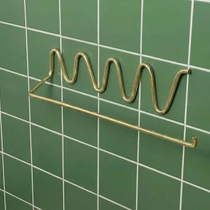 May include: A brass-coloured towel rail with a wavy design and a straight bar for hanging towels. The rail is mounted on a green tiled wall. The design is modern and minimalist.