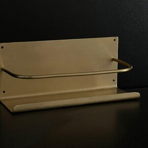May include: A gold-colored metal shelf with a curved bar for hanging items. The shelf has a rectangular backplate with two mounting holes and a small lip at the bottom. The design is minimalist and modern.