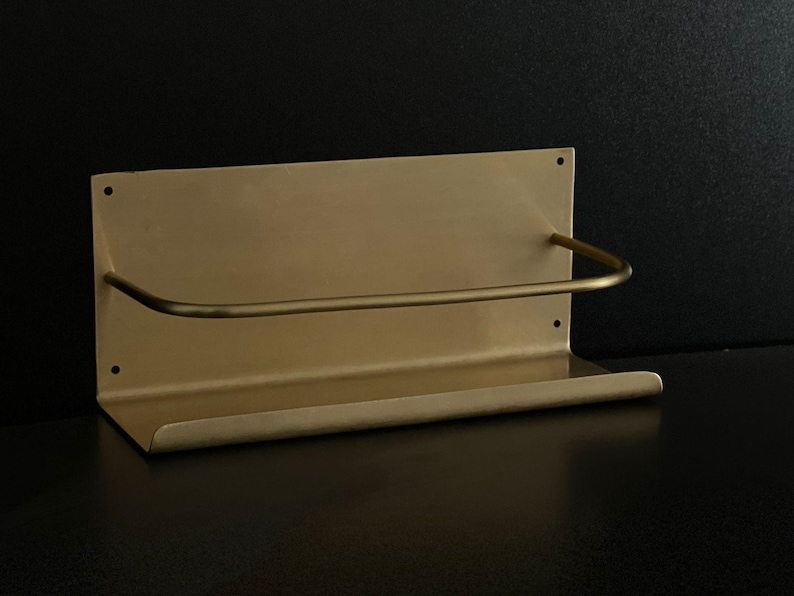 May include: A gold-colored metal shelf with a curved bar for hanging items. The shelf has a rectangular backplate with two mounting holes and a small lip at the bottom. The design is minimalist and modern.