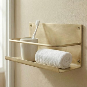 Puede incluir: Estante de pared de color latón con dos niveles. El estante superior contiene una taza de cerámica blanca con un cepillo de dientes. El estante inferior tiene una toalla blanca enrollada. Almacenamiento de baño.