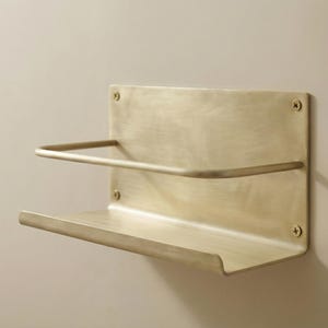 May include: A wall-mounted shelf made of brushed brass. The shelf has a rectangular backplate with a curved top and bottom shelf. A curved bar is above the bottom shelf. Four screws secure the shelf to the wall.
