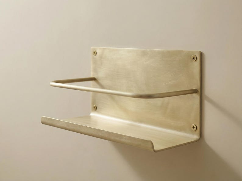May include: A wall-mounted shelf made of brushed brass. The shelf has a rectangular backplate with a curved top and bottom shelf. A curved bar is above the bottom shelf. Four screws secure the shelf to the wall.