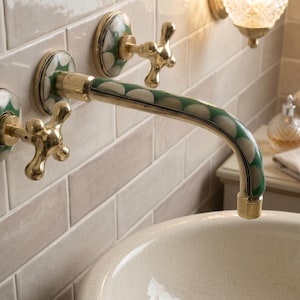 May include: A wall-mounted bathroom faucet featuring a polished brass finish with decorative green and white circular patterns on the handles and the curved spout. The faucet is installed against a light-colored rectangular tiled wall.