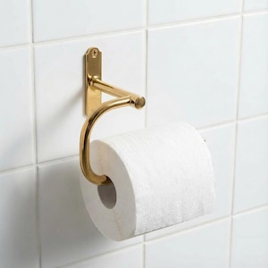 以下が含まれることがあります： 白いタイルの壁に取り付けられた、金色の金属製トイレットペーパーホルダー。白いトイレットペーパーのロールを保持するための湾曲したアームがあります。シンプルでありながらエレガントなバスルームアクセサリーです。